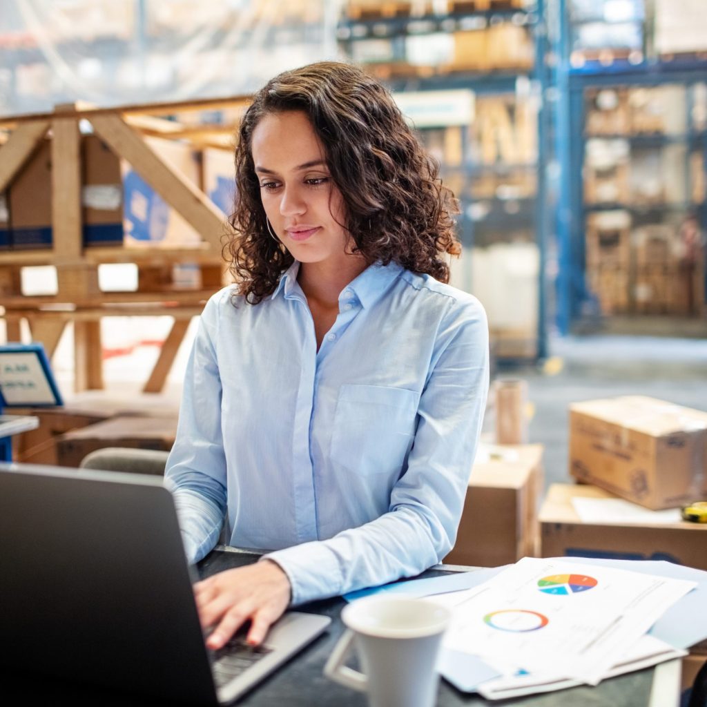 Woman looking busy working on laptop at a distribution warehouse. Businesswoman using laptop to update the stocks at warehouse.