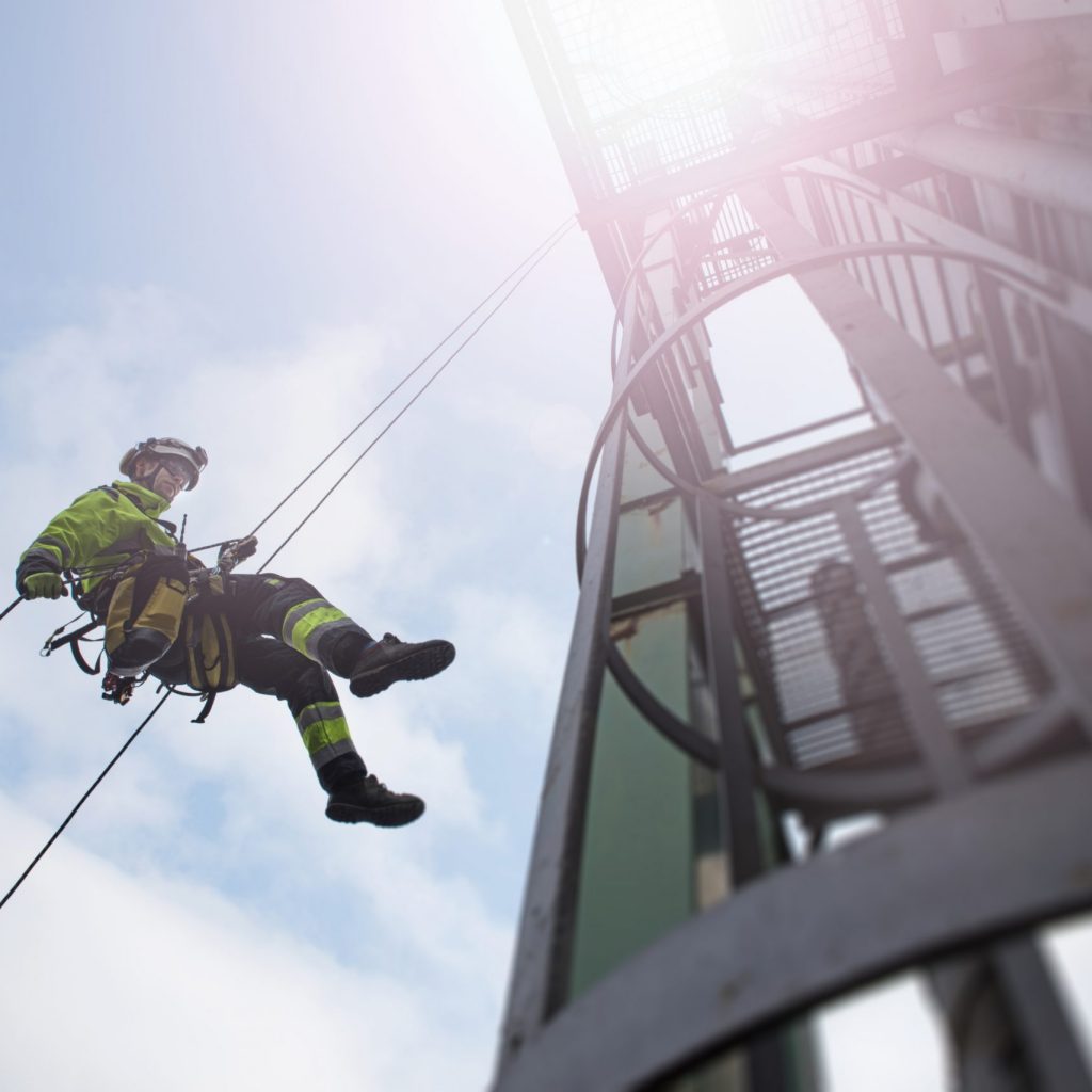 View from bottom on rope access technicians abseil from tower high up with sun behind