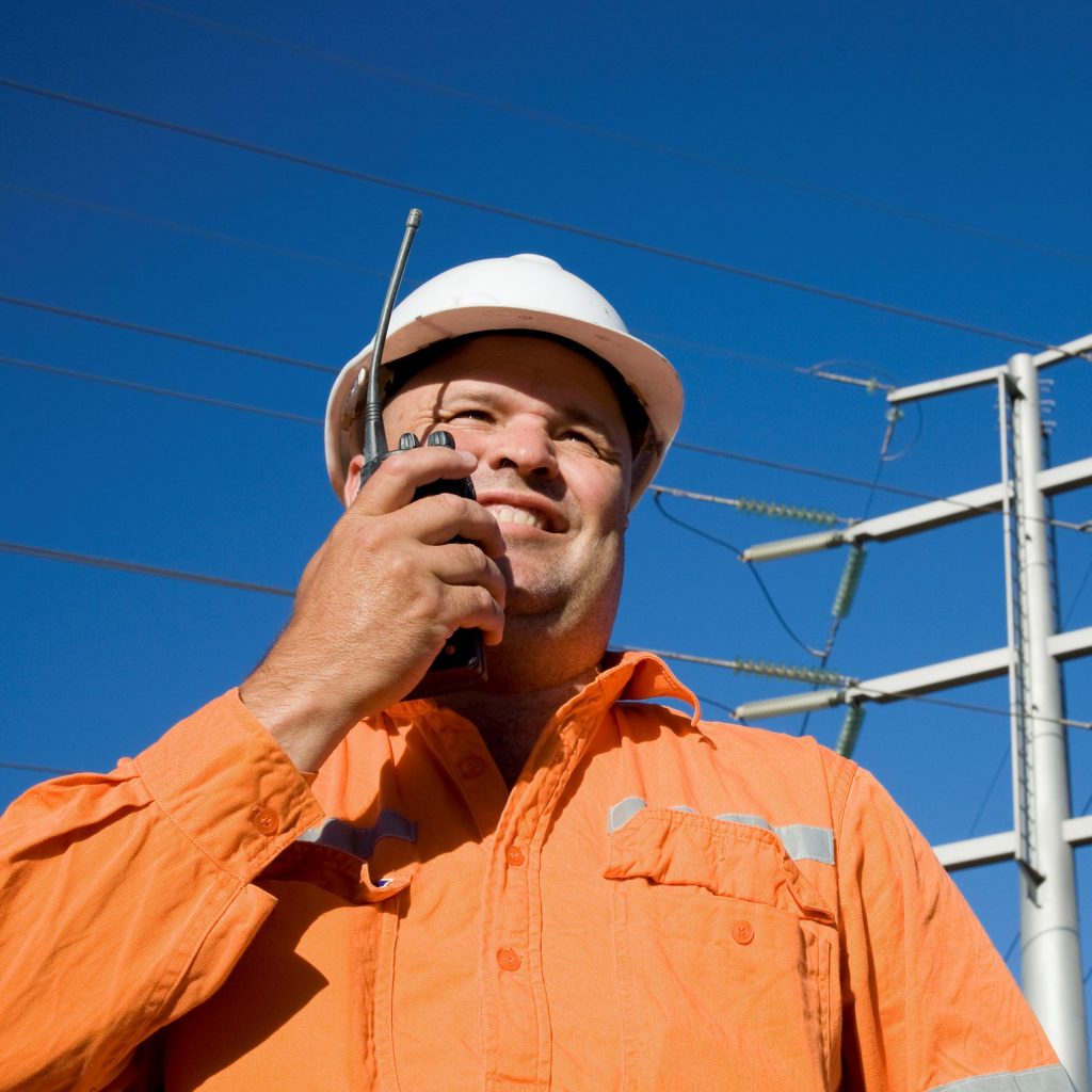 A worker on radio with power lines in background. Could be an Electrician or other Electrical worker in an engineering field. A professional attitude displayed in an industrial environment.