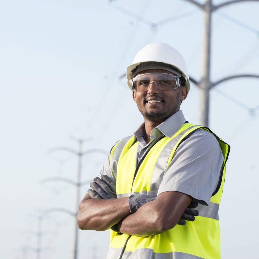 Portrait of manual worker in Personal protective equipment (PPE) standing under high voltage transmission lines against tubular transmission towers.