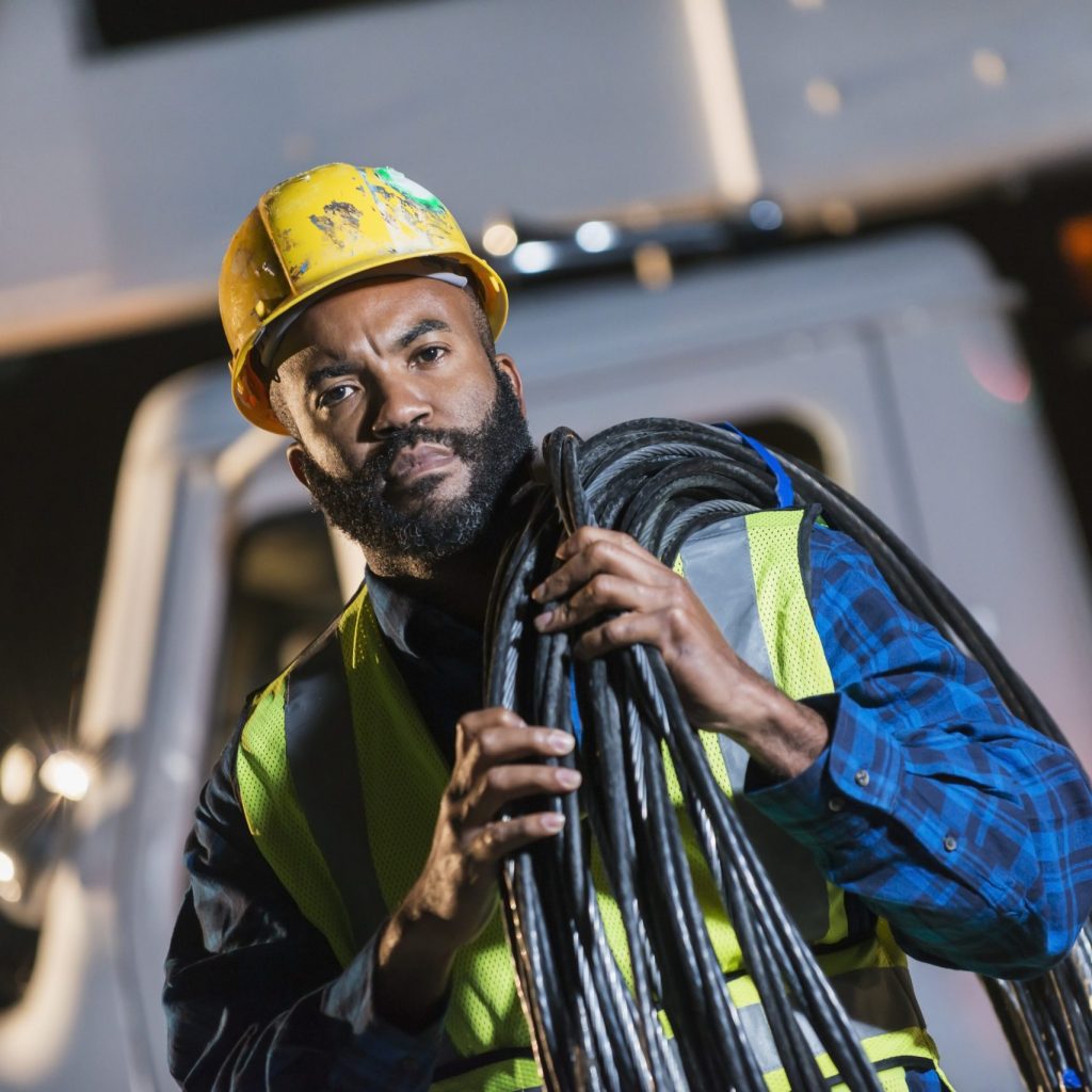 An African American man wearing a hard hat and safety vest, standing in front of a bucket truck or cherry picker, carrying cables on his shoulder. He is a construction worker, utility worker or engineer.