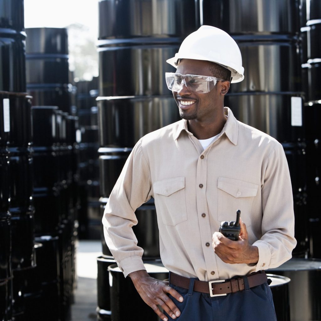 Young African American man, 20s, working at chemical plant, standing by steel drums with walkie-talkie.