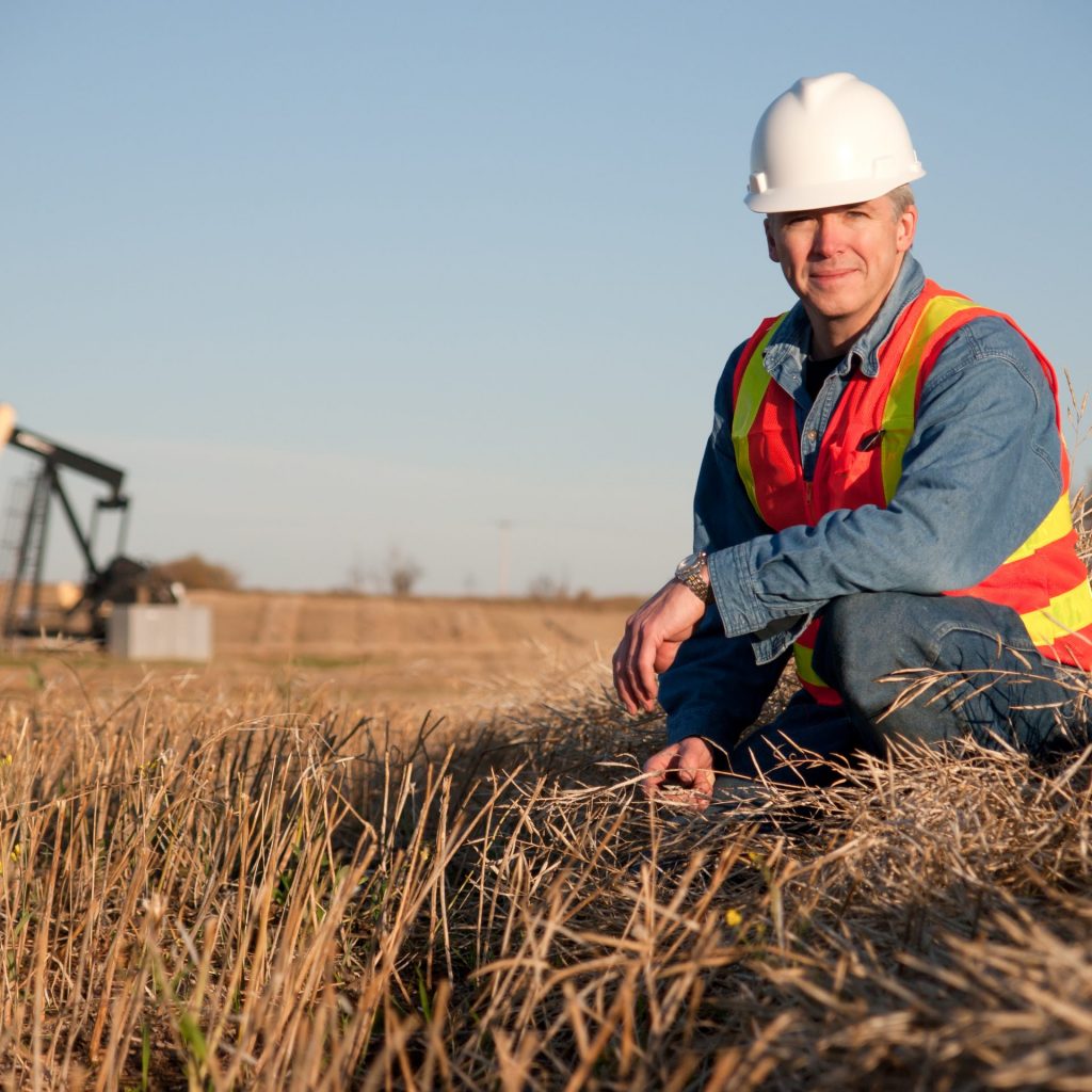 Tridon Communications Mission and Values in Alberta Oil worker engineer in safety gear inspects the soil near an oil well pumpjack. Alberta Canada in the fall.