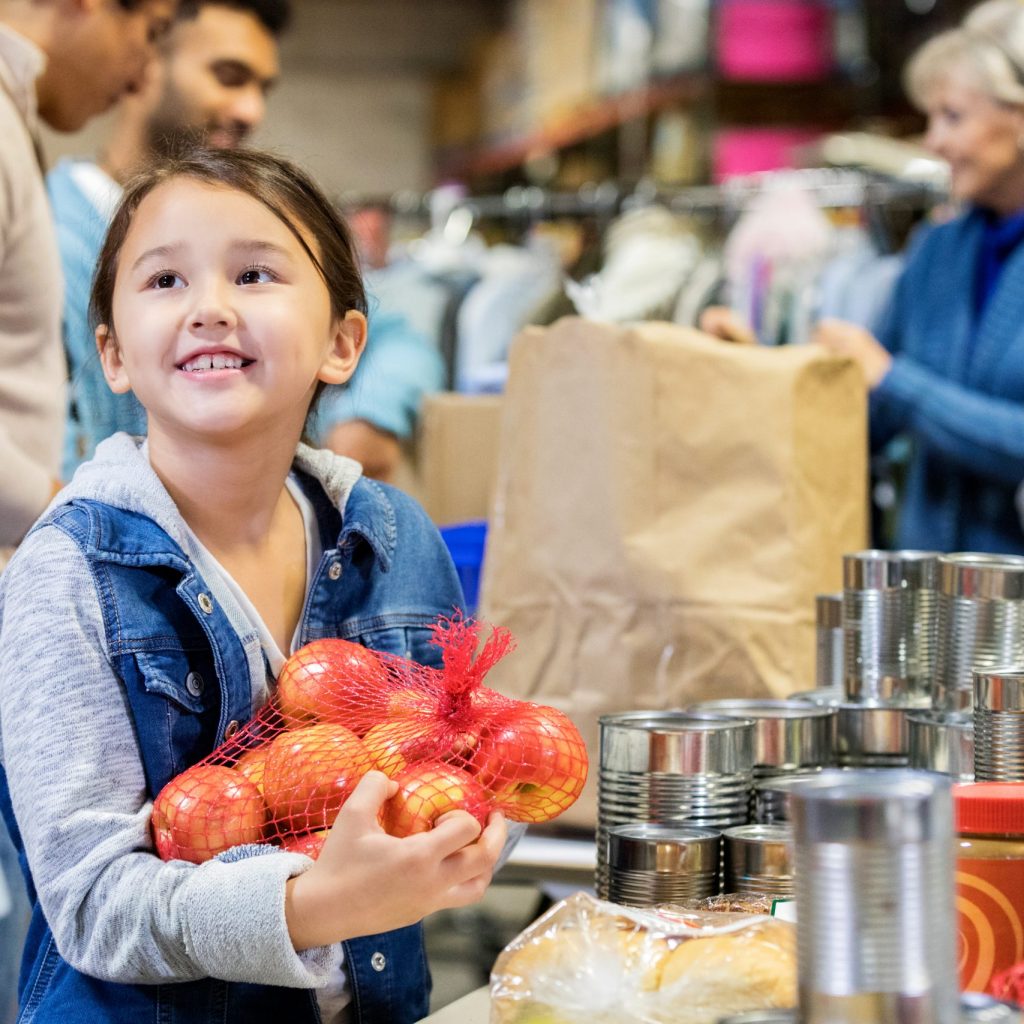 Happy little girl holds a bag of apples while volunteering with her family in a community food bank.